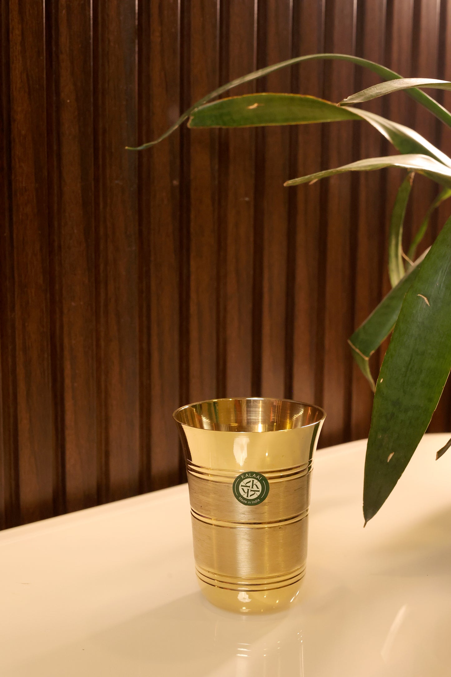Kanchan brass drinking glass on white marble table with wooden panel background